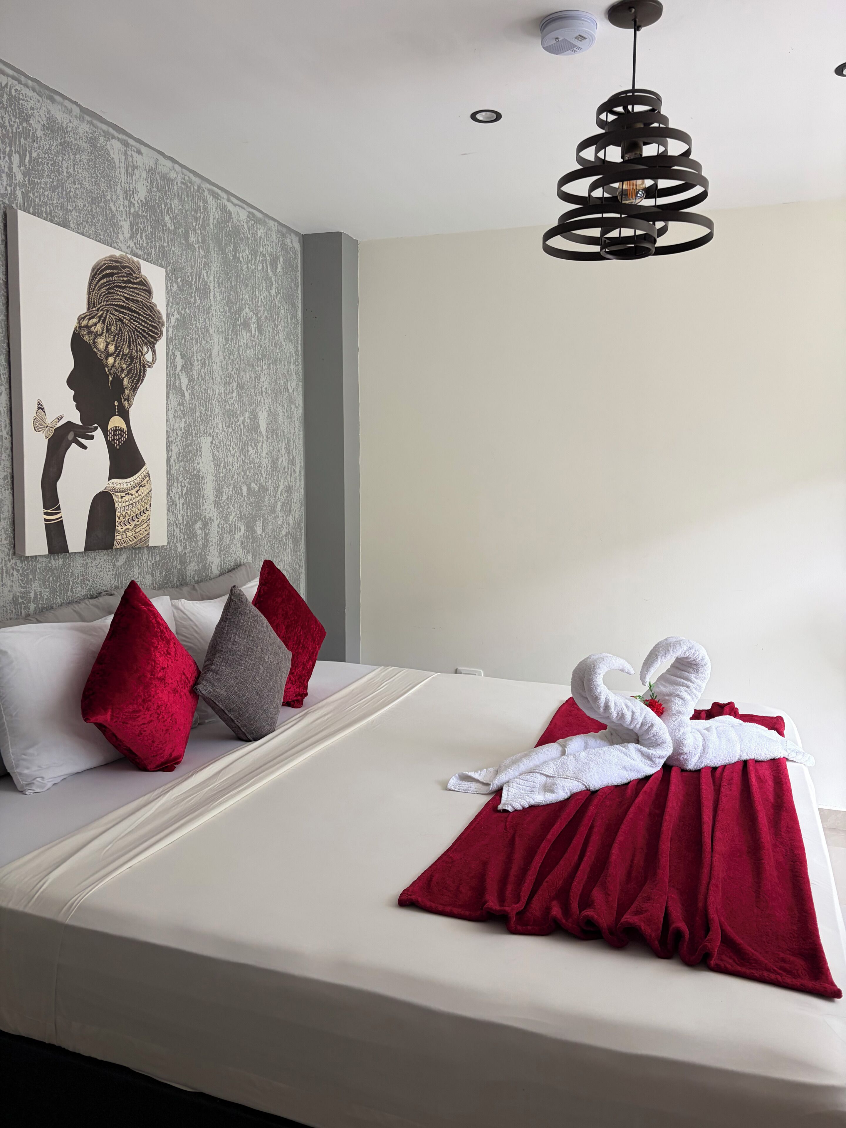 A hotel room with a bed, pillows, a towel swan decoration, a wall art, and a modern ceiling light.