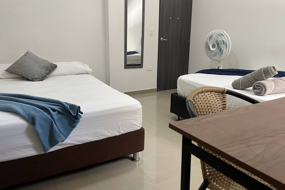 Une chambre d’hôtel avec deux lits, une table, une chaise, un ventilateur et des serviettes.