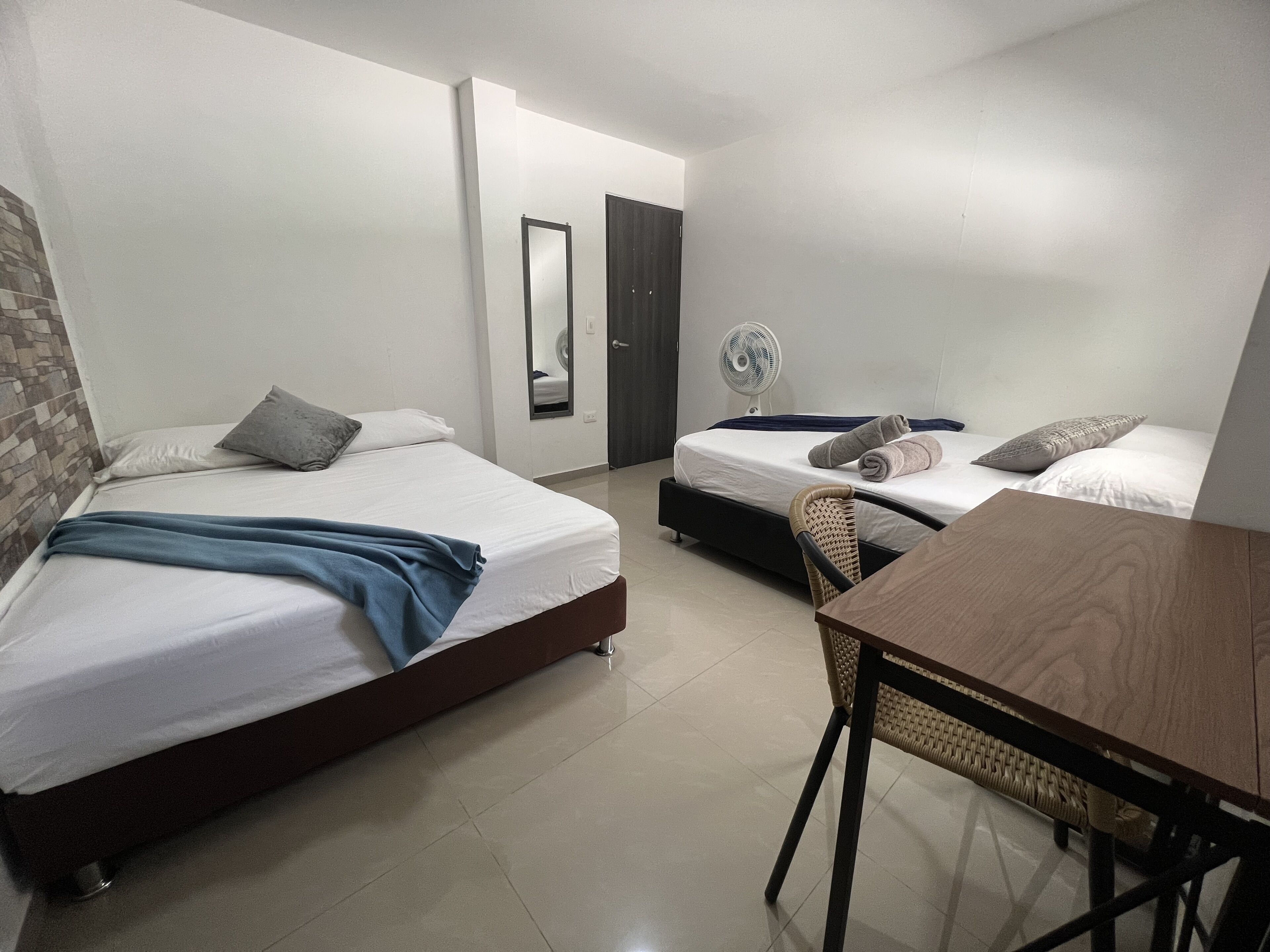 A hotel room with two single beds, a dining table, a chair, a fan, and a mirror.