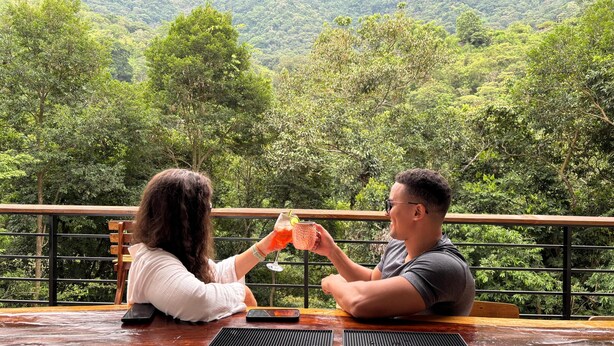 Deux personnes sont assises à une table en bois, devant laquelle s’étend un paysage de montagne boisée.