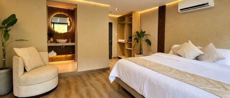 A modern hotel room with a bed, a seating area, a vanity with a mirror, and a built-in air conditioning unit.