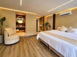 A modern hotel room with a bed, a seating area, a vanity with a mirror, and a built-in air conditioning unit.