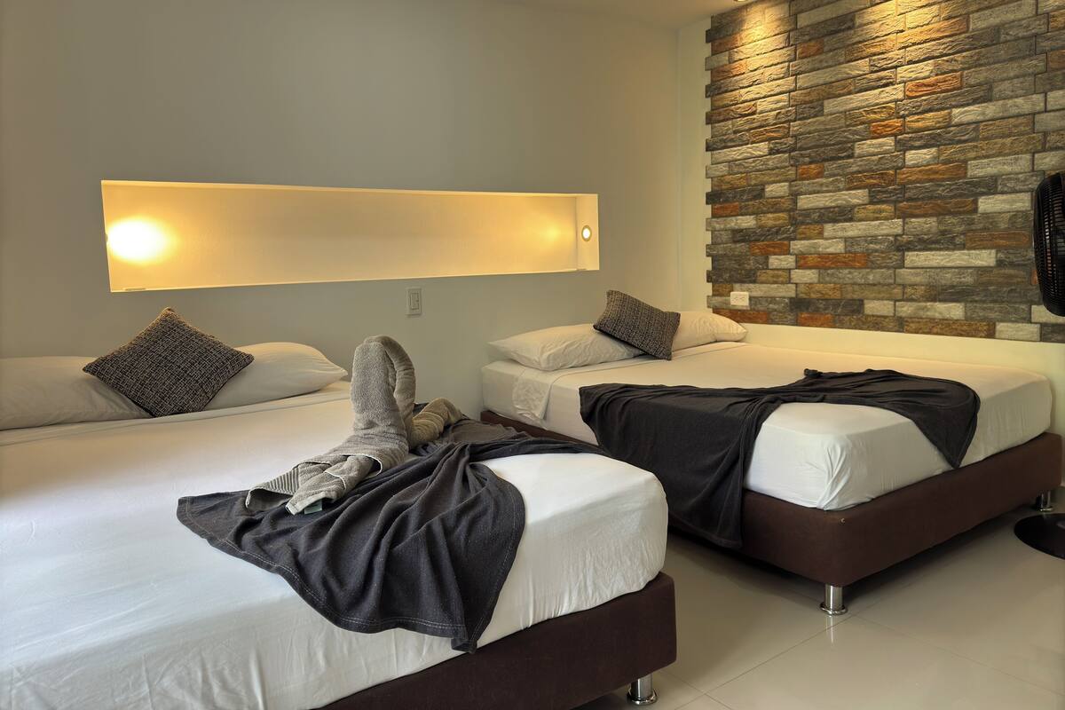 Une chambre d’hôtel avec deux lits, un mur en pierre, une lampe fixée au mur et un ventilateur de plafond.