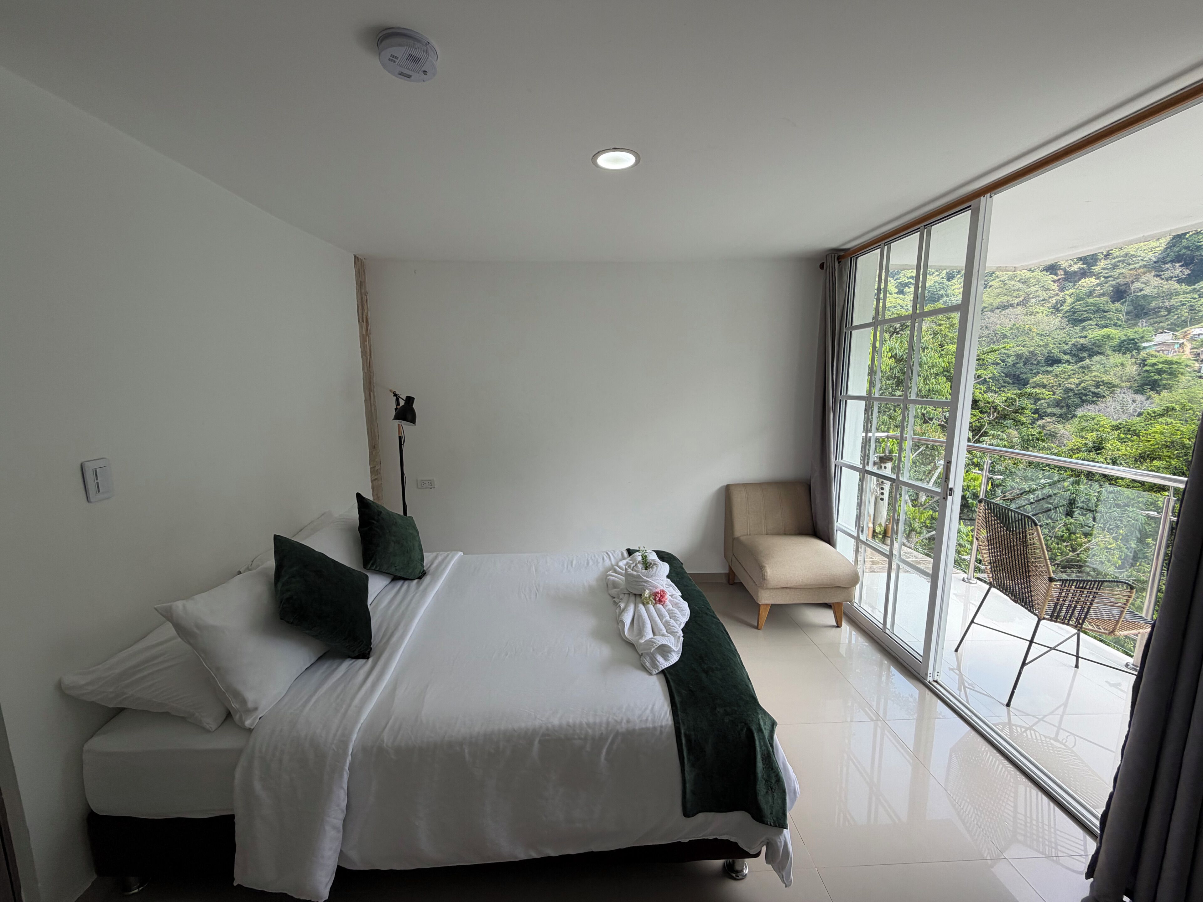 A bedroom with a bed, a chair, a window with a view of greenery, and a ceiling light.