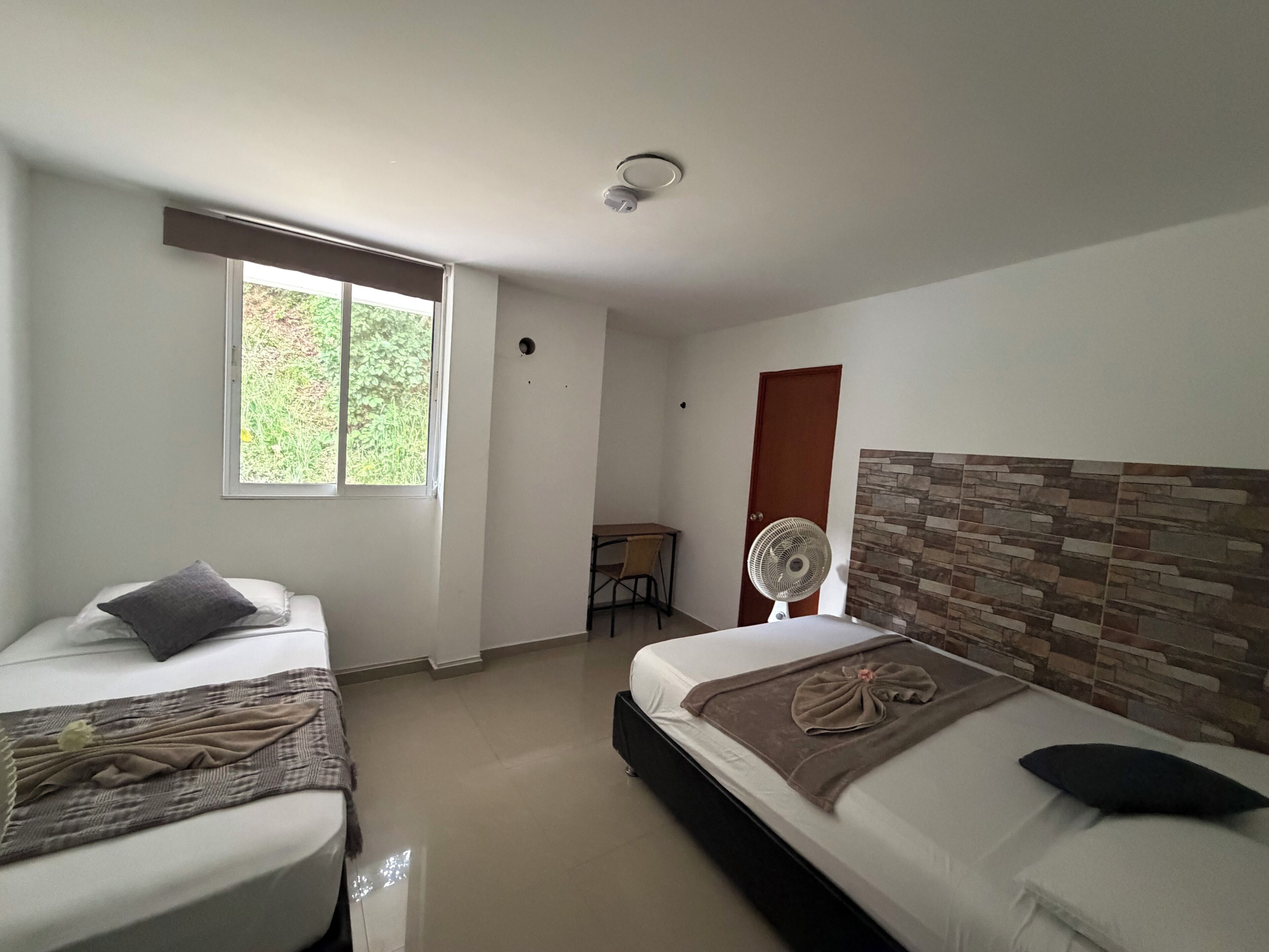 A hotel room with two beds, a window, a fan, a small table, and a door.