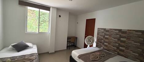 A hotel room with two beds, a window, a fan, a small table, and a door.