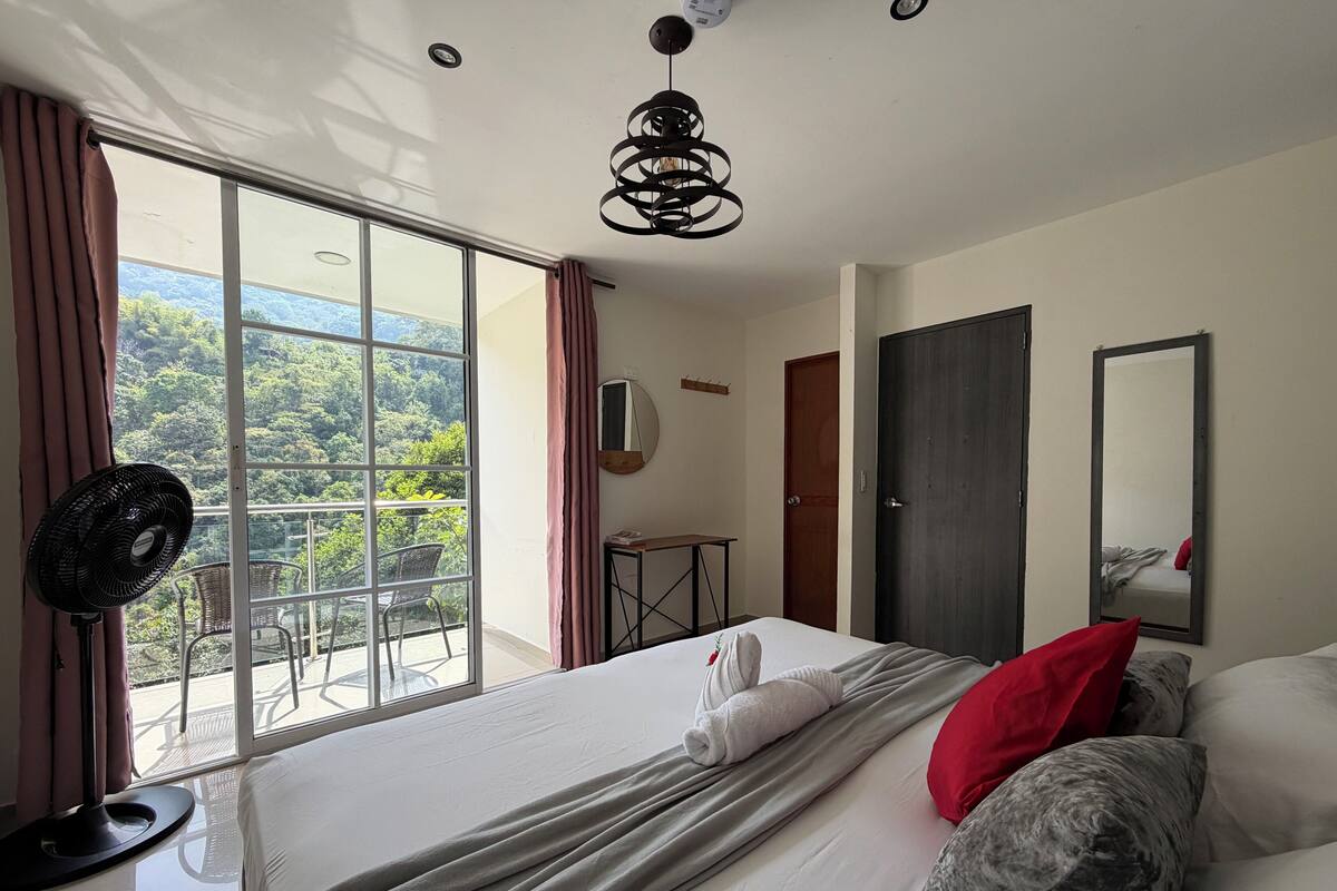 Une chambre d’hôtel avec un grand lit, un ventilateur sur pied, un miroir et une vue sur la verdure par la fenêtre.