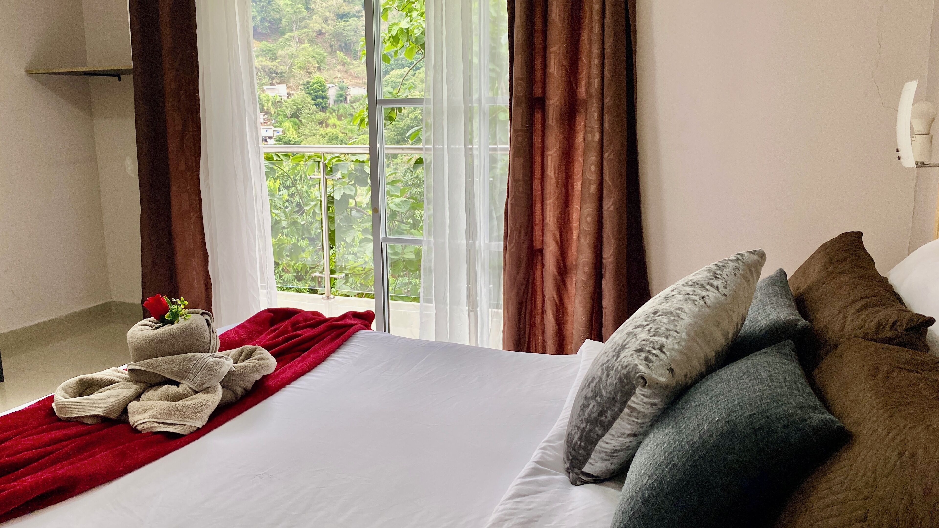 Une chambre d’hôtel avec un grand lit, un plaid rouge et une vue sur la verdure par la fenêtre.
