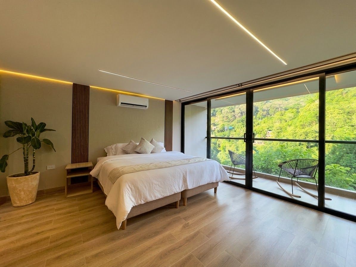 Una camera da letto moderna con un letto grande, una pianta in vaso e vista sul verde attraverso ampie finestre.