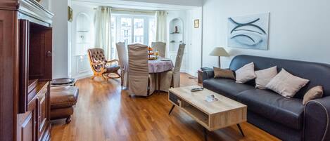 A living room with a sofa, a coffee table, a dining area with chairs, and a wooden cabinet.