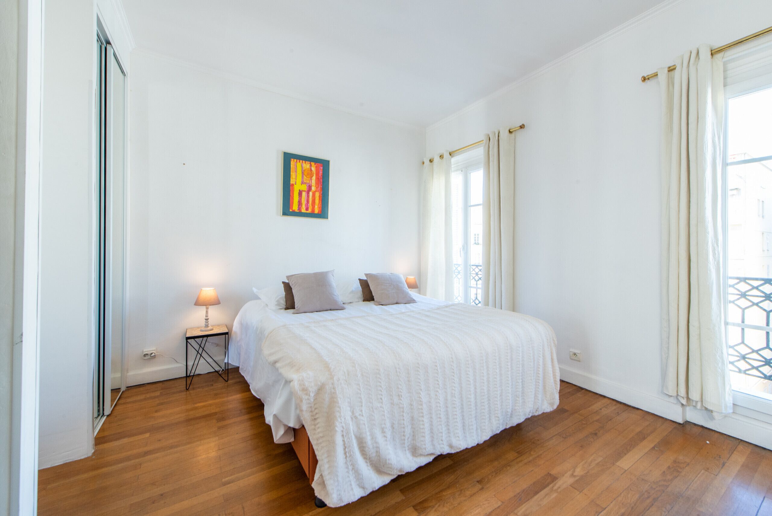 A bedroom with a bed, a nightstand, a lamp, a painting, and a window with curtains.
