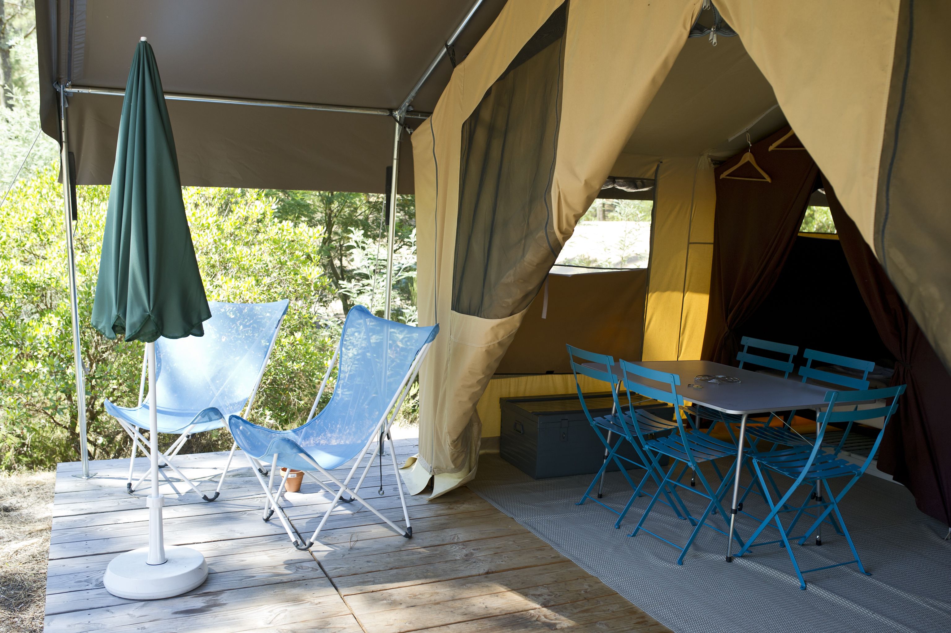A tent with a table and chairs, a green umbrella, and a blue folding chair on a wooden deck.