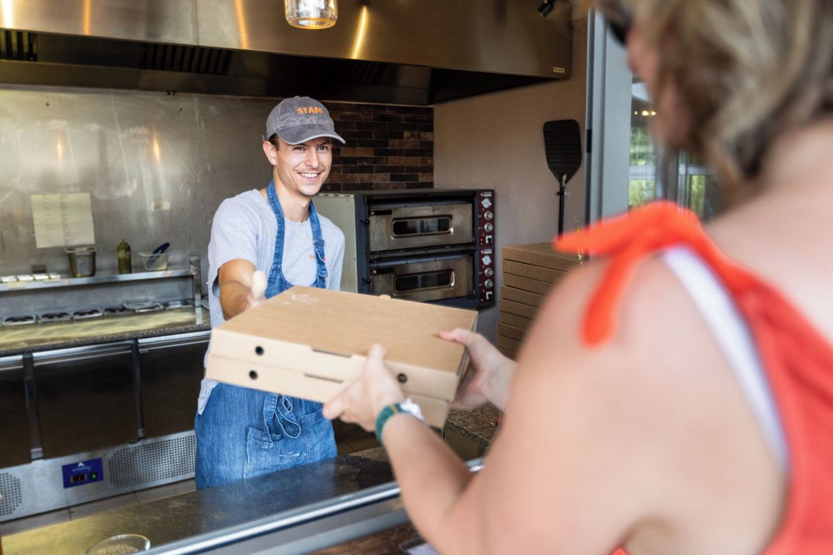 A person handing over a pizza box to another person at a counter with a brick wall and a pizza oven in the background.