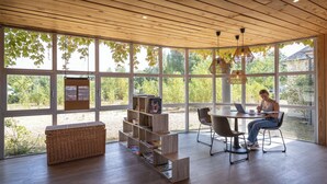 A modern room with large windows, a wooden ceiling, a bookshelf, a table with chairs, and a laptop.