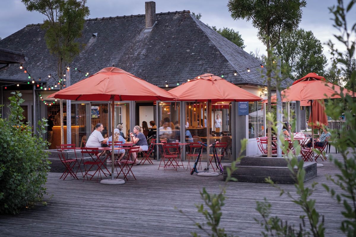 Un espace extérieur aménagé pour les repas, avec des parasols rouges et des tables, entouré d’une terrasse en bois et d’arbres.