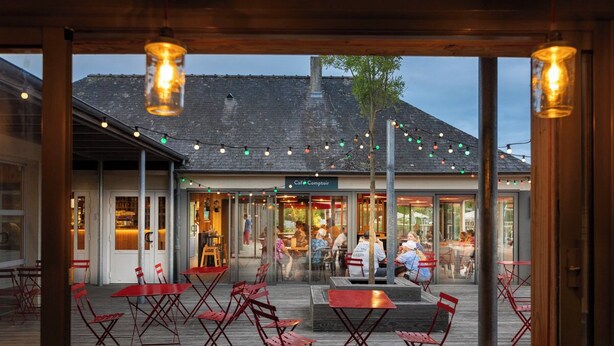 An outdoor café with red tables and chairs, a wooden deck, and a building with a sign reading "Café Comptoir" in the background.