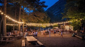 An outdoor dining area with picnic tables and string lights, surrounded by trees and a mountain backdrop.