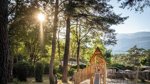 A playground with a wooden climbing structure and a slide, surrounded by trees and a view of distant mountains.