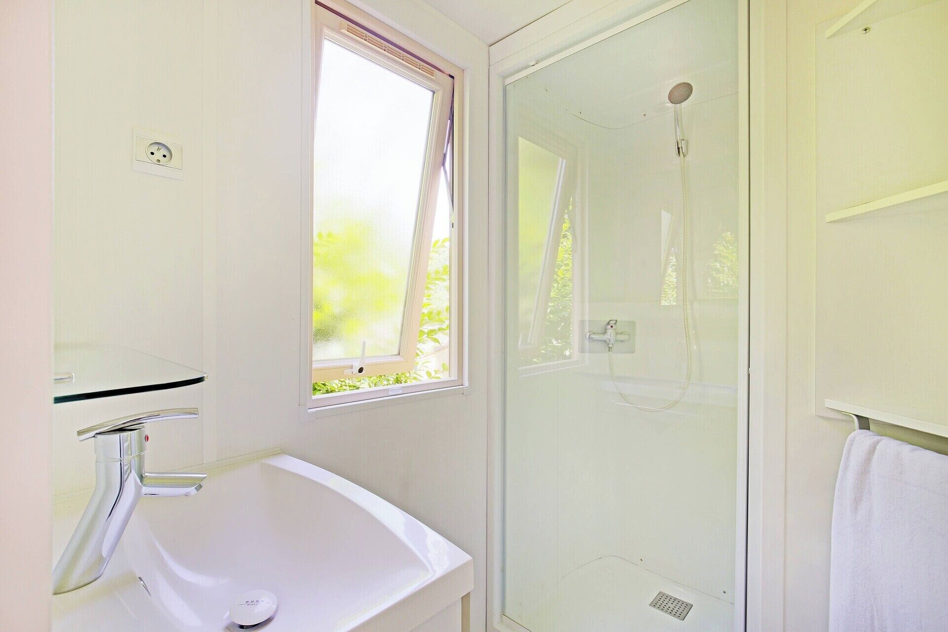 Un baño con cabina de ducha de vidrio, un lavabo blanco y una ventana con vistas a la vegetación.