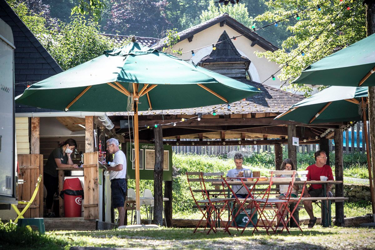 Un café en plein air avec des tables en bois et des parasols verts, entouré de verdure et avec un bâtiment en arrière-plan.