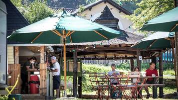 Un café en plein air avec des tables en bois et des parasols verts, entouré de verdure et avec un bâtiment en arrière-plan.