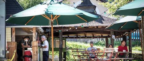 An outdoor café with wooden tables and green umbrellas, surrounded by greenery and a building in the background.