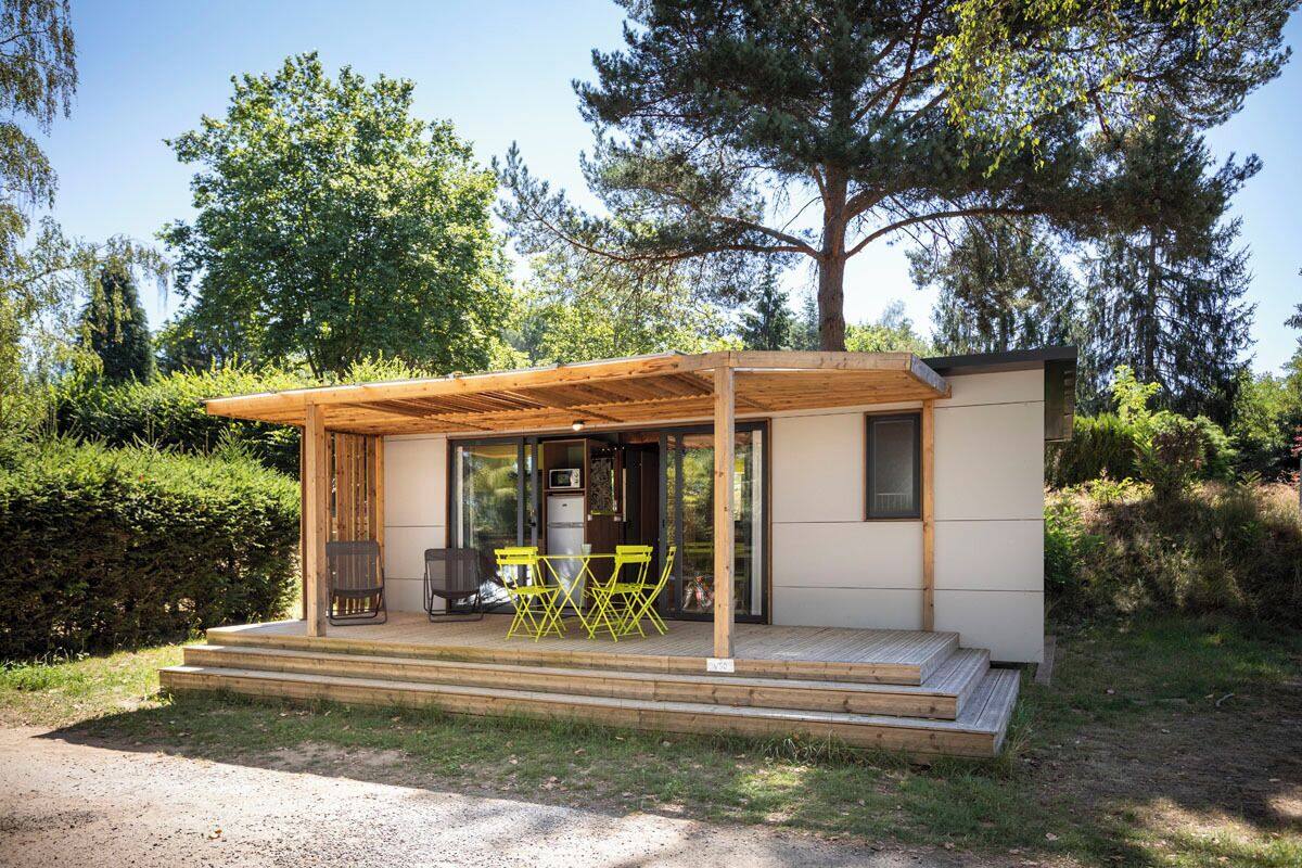 Une petite cabane moderne avec un porche en bois, un coin repas avec des chaises jaunes et une cuisine visible à travers les portes vitrées.