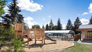 Un parque infantil con estructuras de madera para escalar y un tobogán, rodeado de árboles y un edificio con techo curvo.