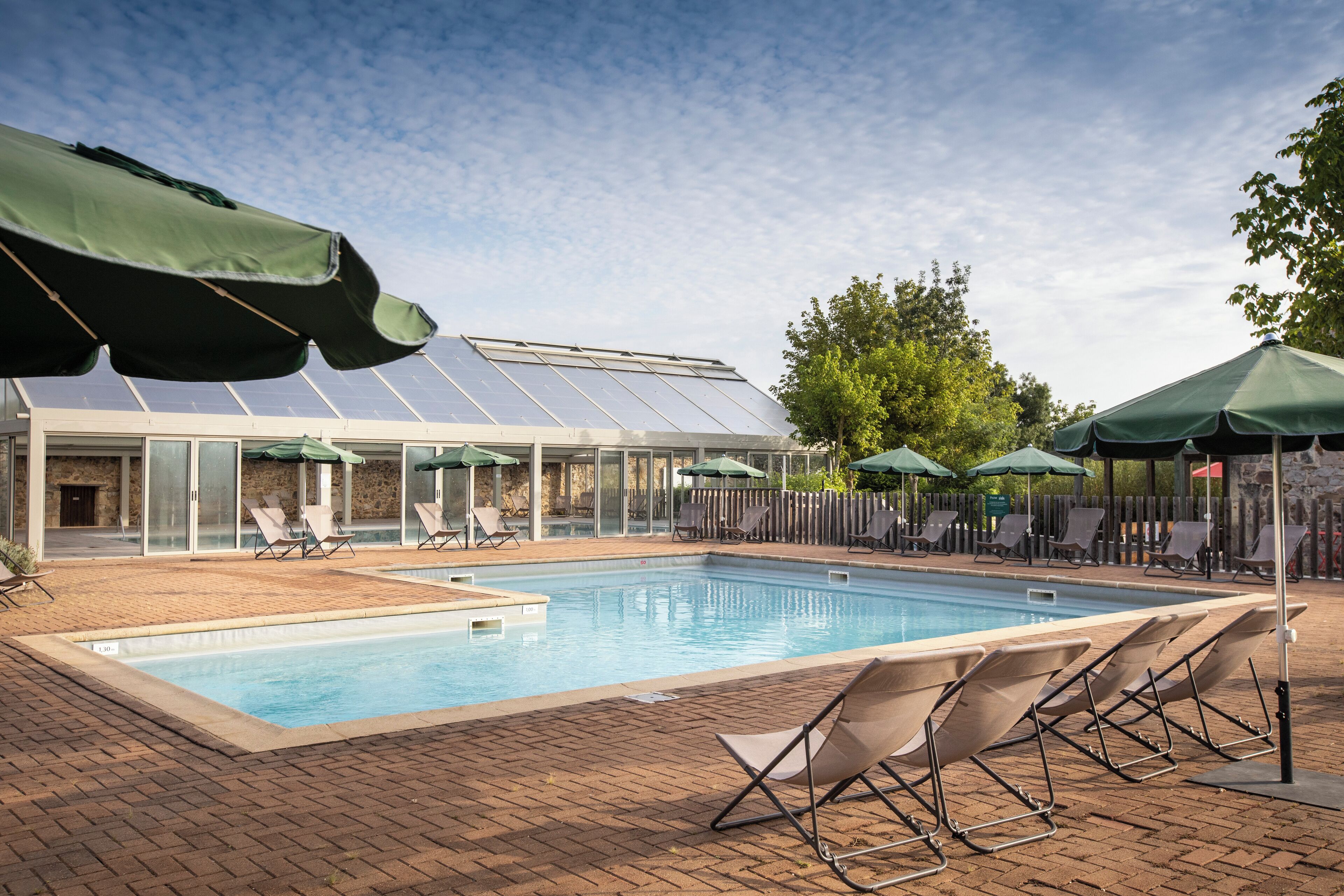 Une piscine entourée de chaises longues et de parasols, elle-même bordée d’une terrasse en briques et d’un bâtiment à panneaux de verre abritant des panneaux solaires.