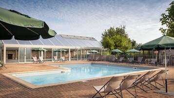 Une piscine entourée de chaises longues et de parasols, elle-même bordée d’une terrasse en briques et d’un bâtiment à panneaux de verre abritant des panneaux solaires.