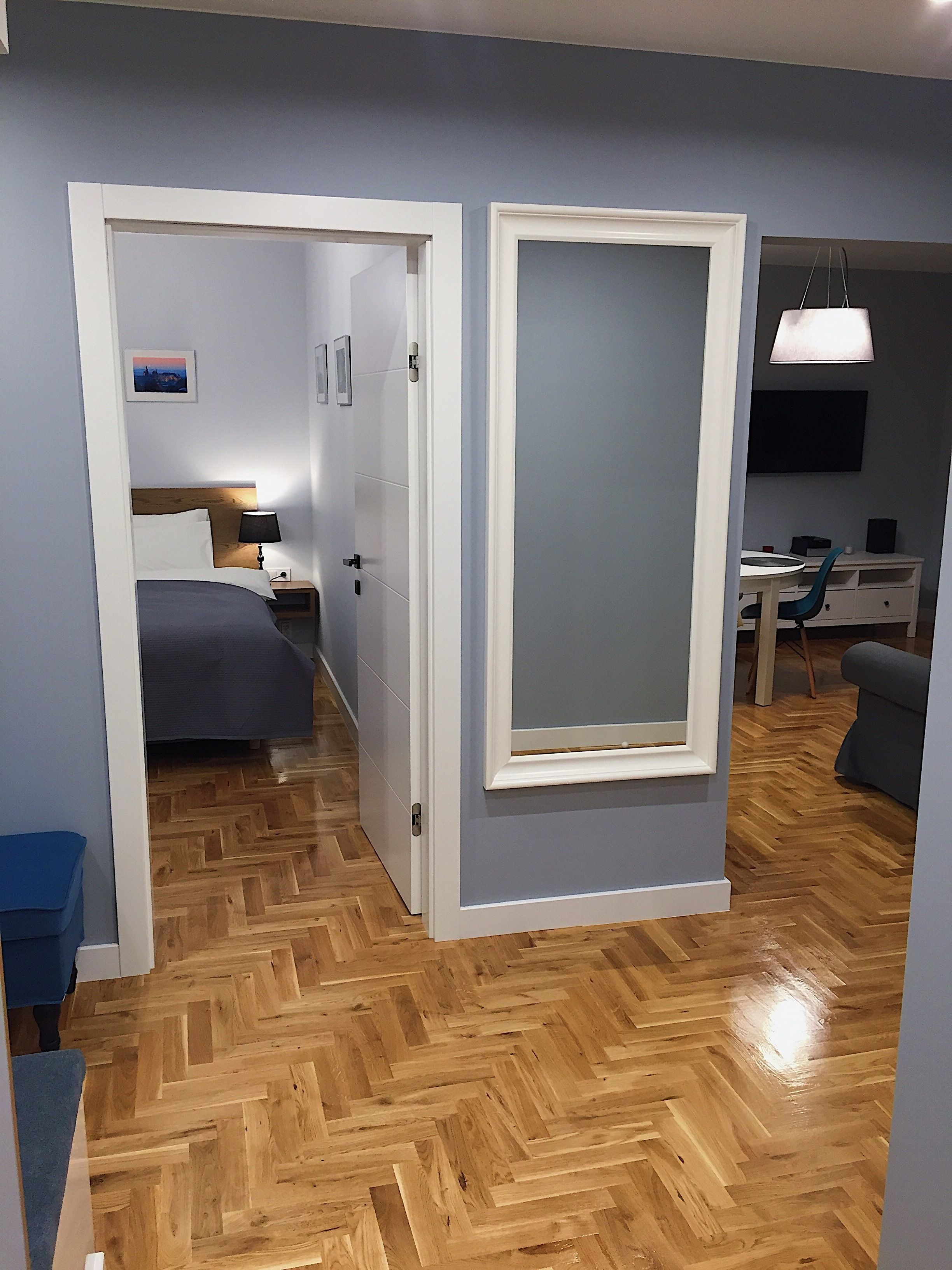 A modern hotel room with a wooden herringbone floor, a bed with a wooden headboard, a desk with a chair, and a TV mounted on the wall.