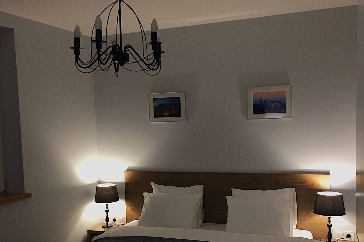 A bedroom with a large bed, two bedside tables with lamps, a chandelier, and two framed pictures on the wall.