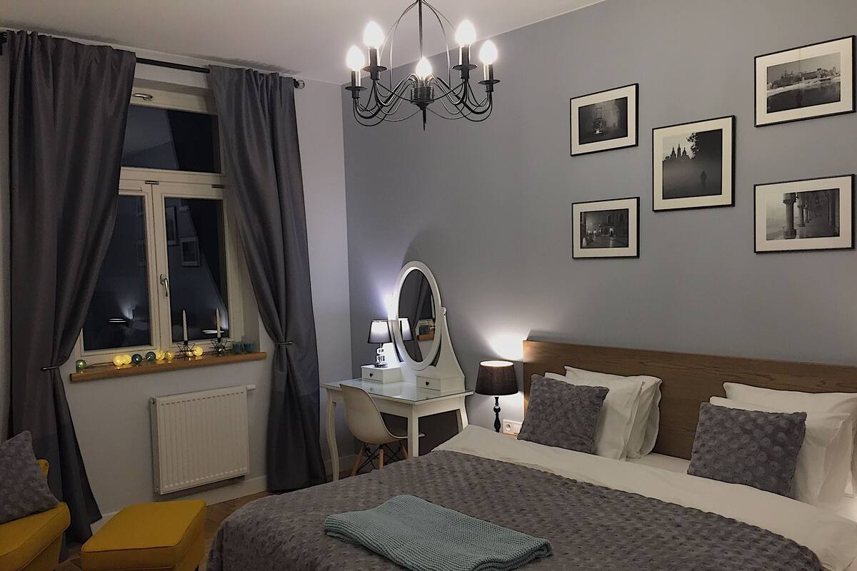 A bedroom with a bed, bedside table, lamp, mirror, and a window with curtains.