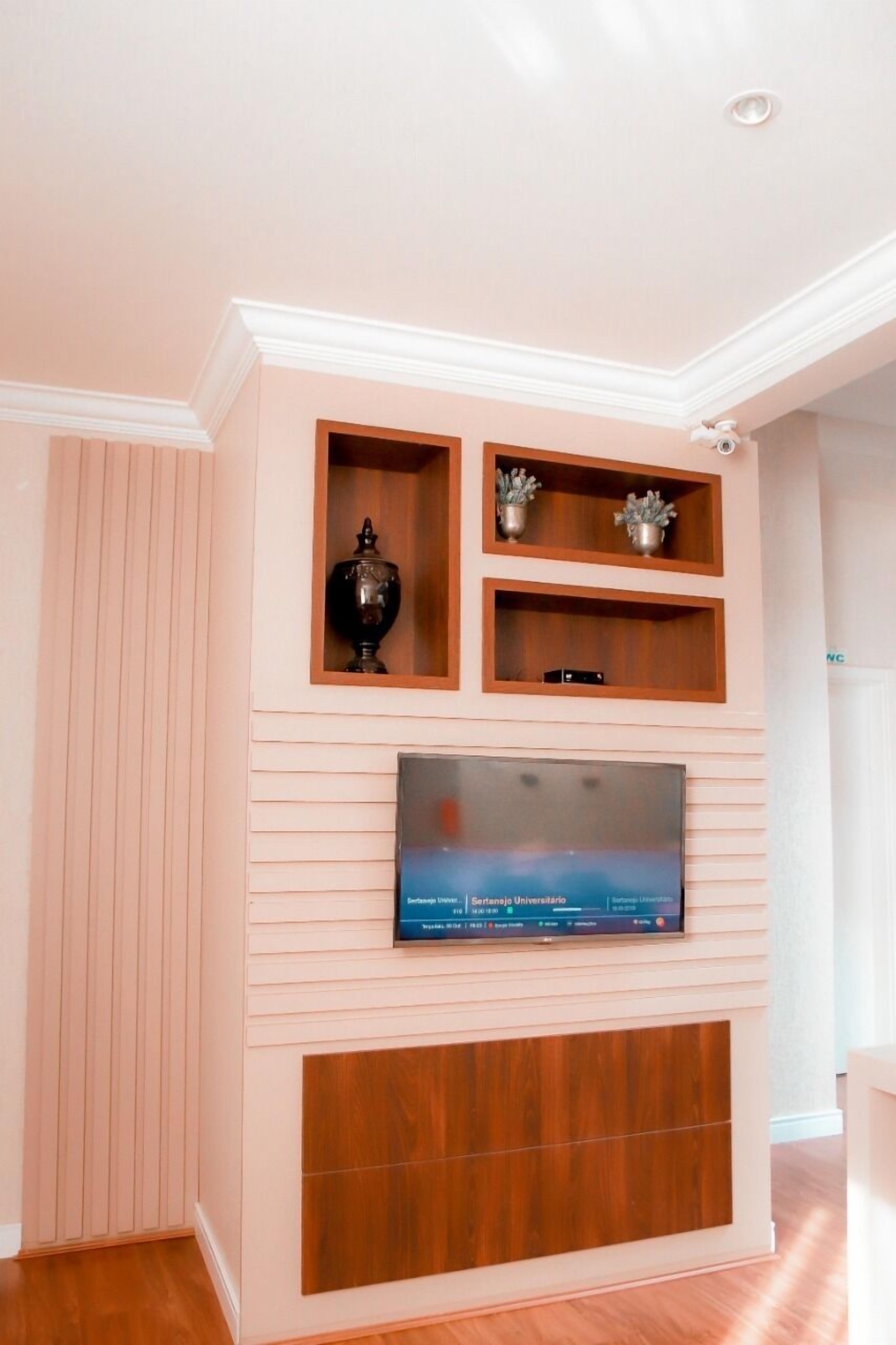 Uma sala de estar com uma televisão de tela plana fixada em um painel de madeira, um vaso decorativo e uma pequena estante com plantas.