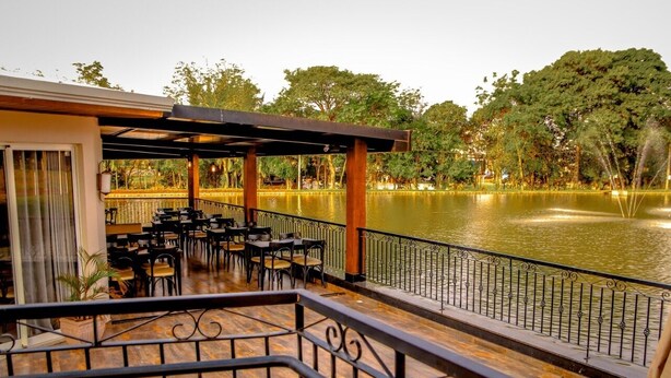 An outdoor dining area with tables and chairs, a wooden deck, and a view of a lake with a fountain. - Mirante Hotel (Itapejara d'Oeste)