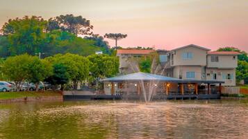 Hotel à beira do lago com uma fonte, rodeado por árvores e um estacionamento.