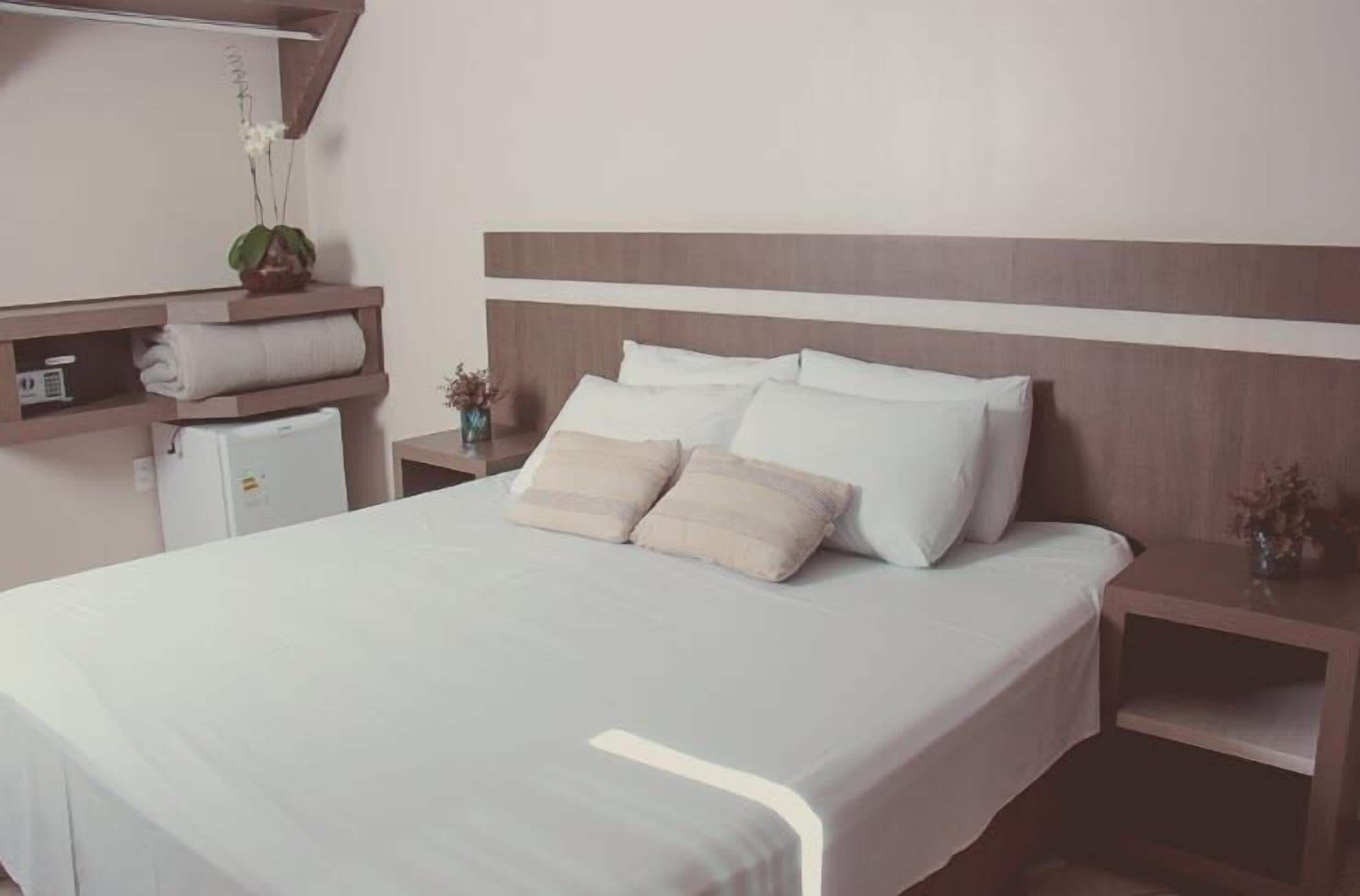 Una cama bien tendida con ropa de cama blanca y dos almohadas, una mesita de noche con un florero y una pequeña estantería con una toalla doblada y una planta.