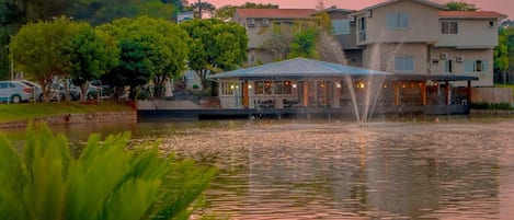 Un hôtel situé au bord d’un lac, doté d’une fontaine, entouré d’arbres et offrant une vue sur un ciel dégagé.