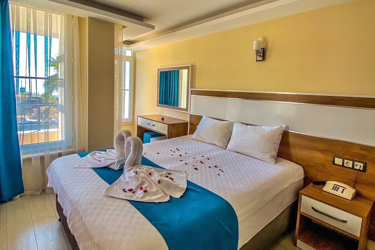 A hotel room with a large bed, a bedside table with a phone, a nightstand, a window with curtains, and a ceiling light.