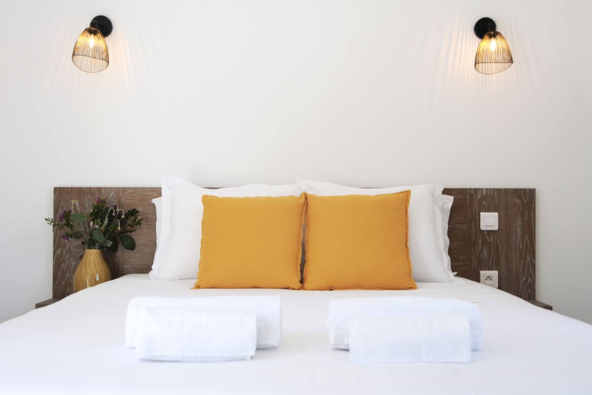 Uma cama bem arrumada com almofadas brancas e mostarda, duas luminárias de cabeceira e um vaso com flores na mesa de apoio.