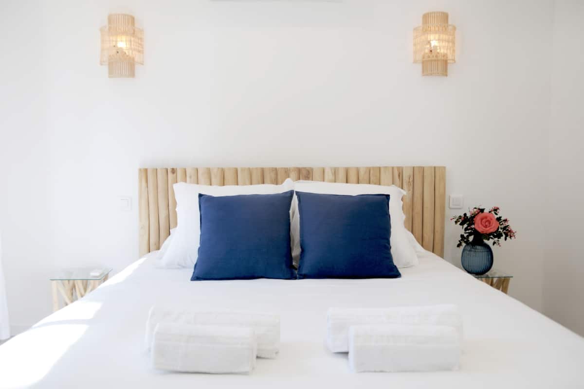 Cama bem arrumada com almofadas brancas e azuis, duas luminárias de cabeceira e um vaso com flores em uma mesa de apoio.