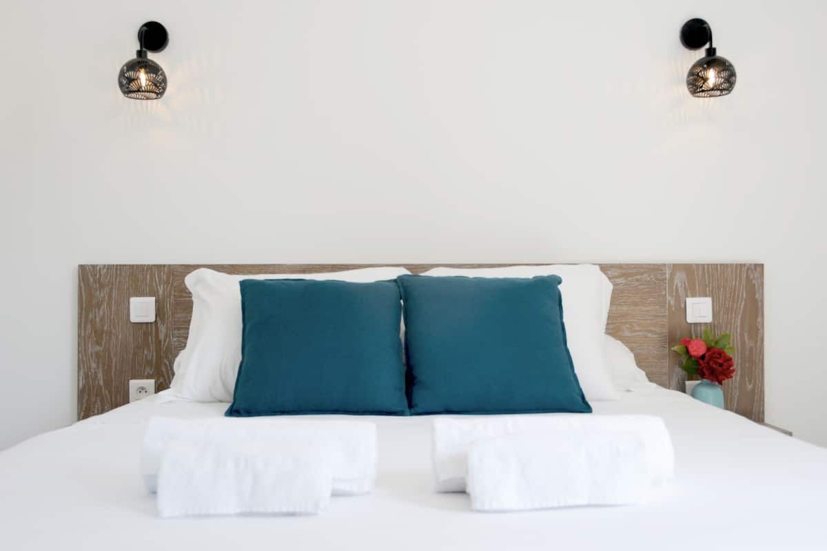 Cama bem arrumada com almofadas brancas e azul-turquesa, cabeceira de madeira e duas luminárias fixadas na parede.