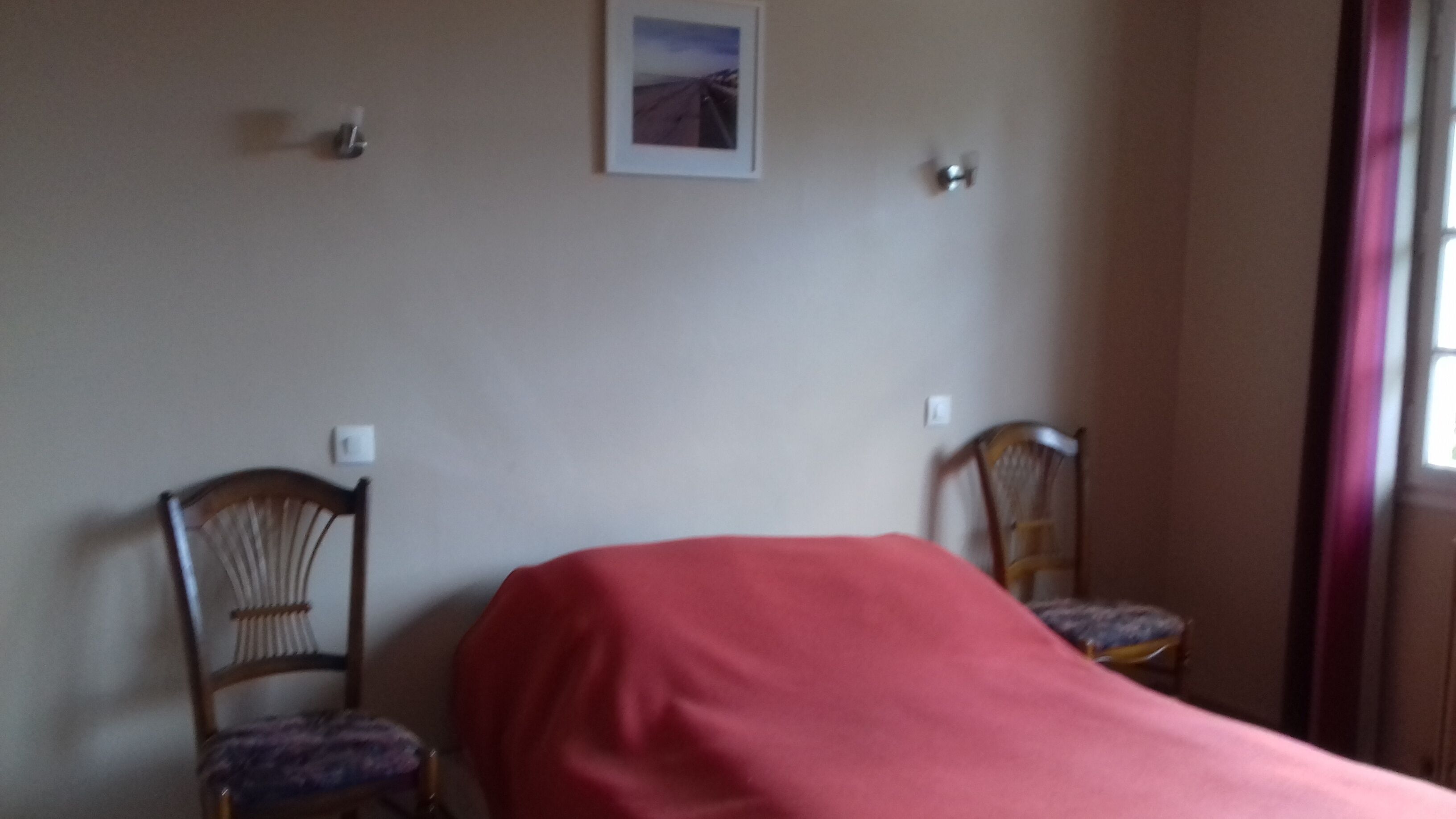 A room with a bed covered in a red blanket, two wooden chairs, a framed picture on the wall, and a window with curtains.