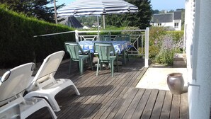 An outdoor patio with a table and chairs under a striped umbrella, surrounded by greenery and a view of houses in the background. - House in Balearic resort at 900 m. from the beach and shops (Jullouville)