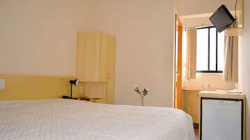 Habitación de hotel con cama, escritorio con lámpara, una mesita con planta y ventana con cortinas.