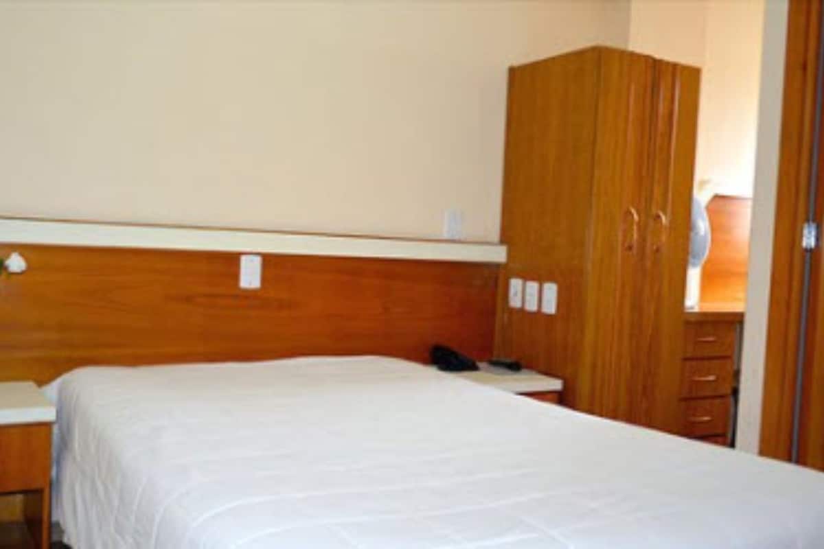 A hotel room with a wooden headboard, a white bedspread, a bedside table with a lamp, and a wardrobe.