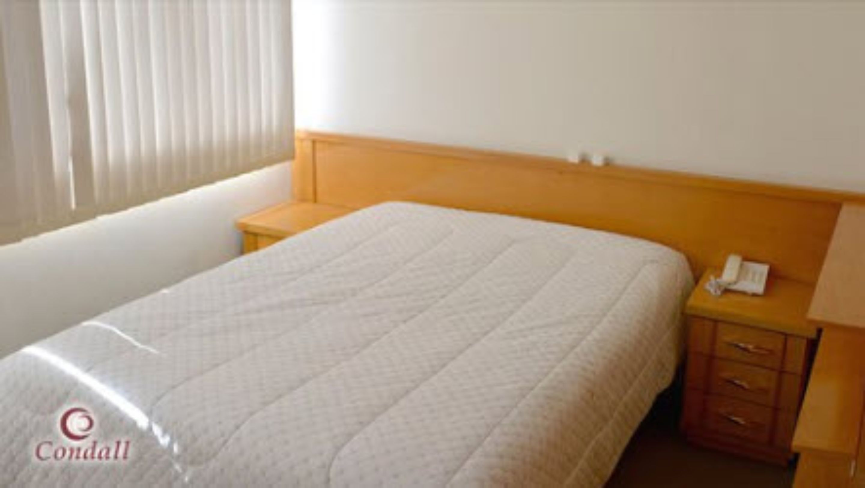 A hotel room with a large bed, a wooden headboard, a bedside table with a phone, and a window with blinds.