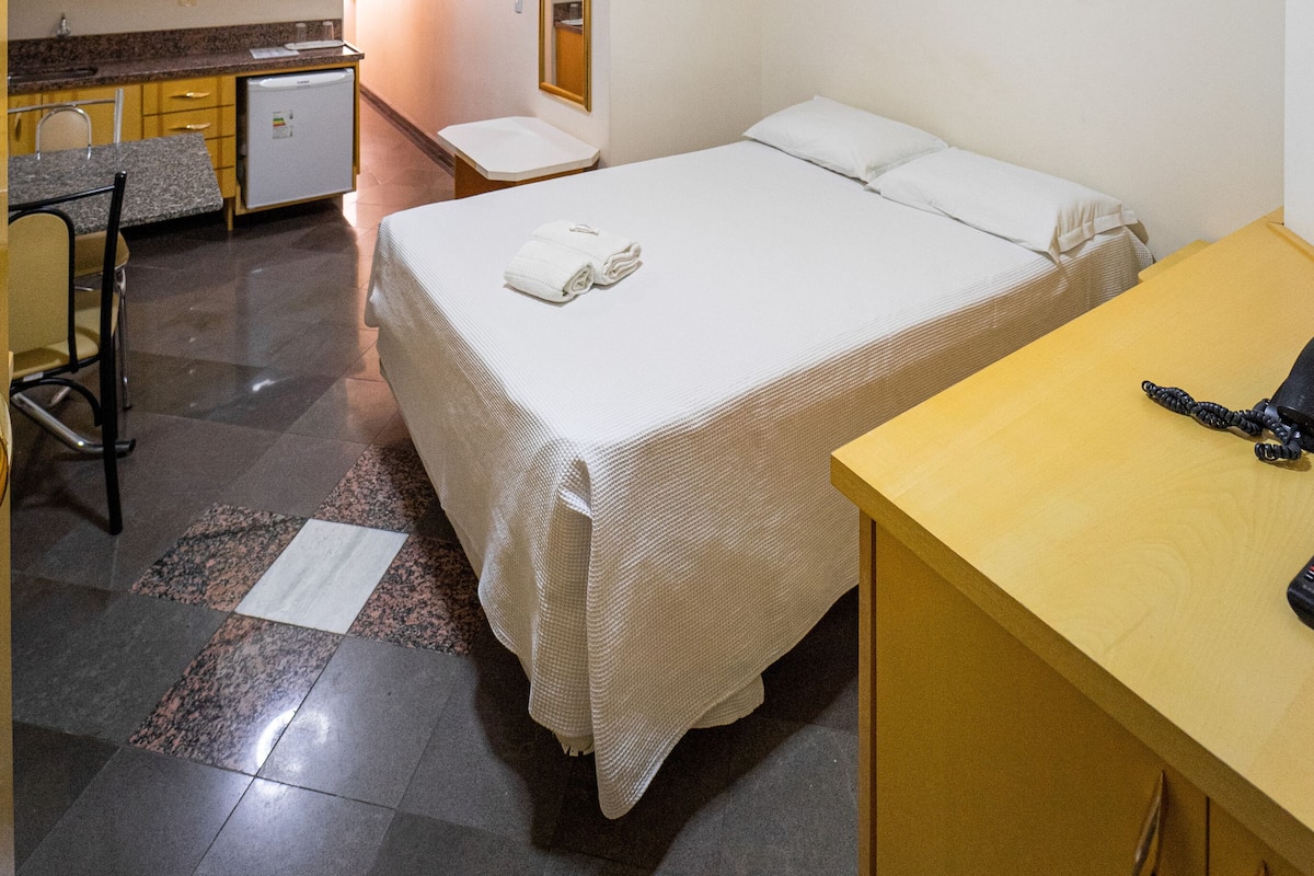 A compact hotel room with a bed, a small desk, a chair, a kitchenette, and a bathroom.
