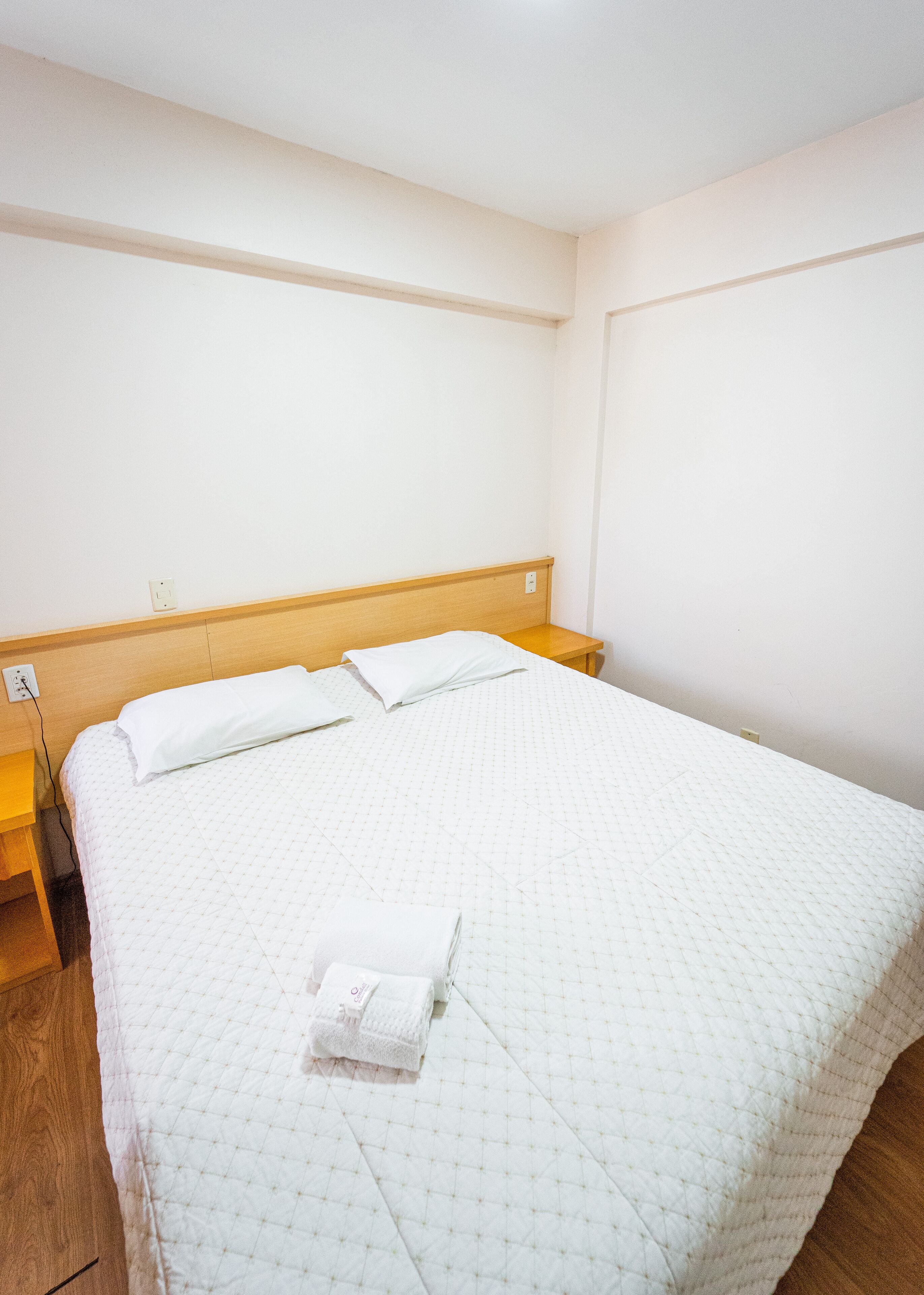 Un letto rifatto con cura, con una coperta bianca trapuntata, due cuscini e un asciugamano bianco piegato.