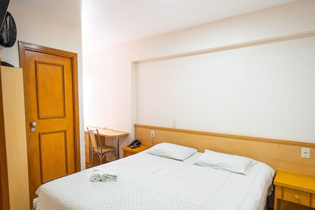 A neatly made bed with a white quilt, a wooden headboard, a bedside table with a lamp, and a wooden wardrobe.